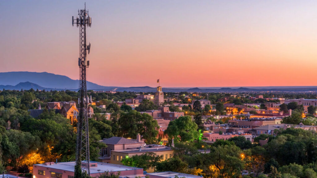 Cell tower stands among buildings and trees at sunset in a small city, with mountains visible in the background under a colorful sky.