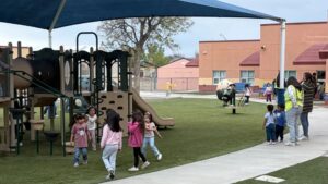 Young children play on a playground and walk on a path under a blue shade canopy, with adults supervising nearby on a grassy area outside a school building.