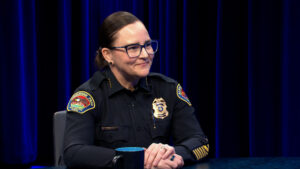 A police officer in uniform sits at a table with a blue mug, smiling, against a dark blue curtain background.