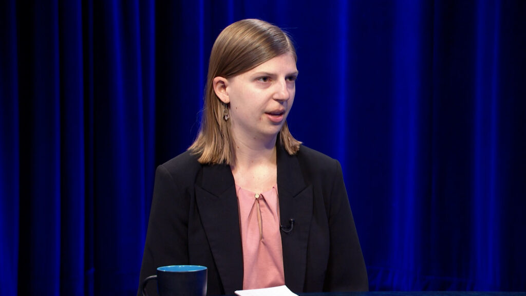 A woman with straight, shoulder-length hair in a black blazer and pink blouse speaks in a studio with blue curtains. A blue mug and papers are on the table in front of her.