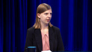 A woman with straight, shoulder-length hair in a black blazer and pink blouse speaks in a studio with blue curtains. A blue mug and papers are on the table in front of her.