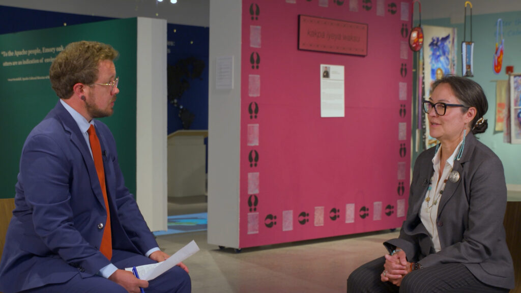 A man in a blue suit and a woman in a black suit are having a seated conversation in a museum or exhibition space with colorful displays in the background.