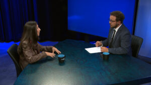Two people sit across from each other at a blue table in a studio, engaged in conversation. One holds a pen and paper; both have mugs in front of them.