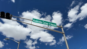 A street sign reading "Avenida Cesar Chavez" mounted on a metal pole, with a background of blue sky and scattered clouds.