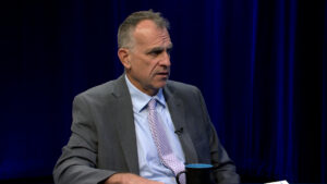 A man in a gray suit and light-colored tie sits at a table with a blue and black mug, speaking against a dark blue background.
