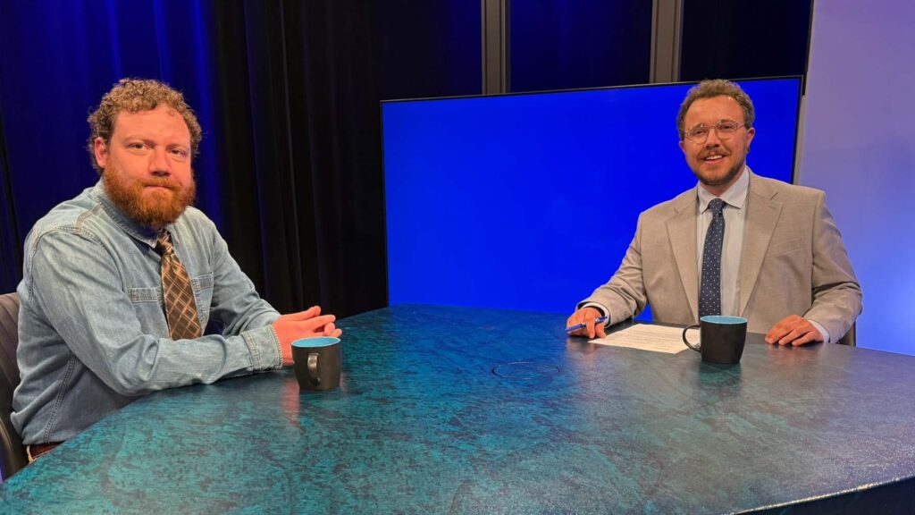 Two men sit at a table with coffee mugs in a studio setting; one wears a denim shirt, the other a suit, with a blank blue screen in the background.