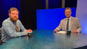 Two men sit at a table with coffee mugs in a studio setting; one wears a denim shirt, the other a suit, with a blank blue screen in the background.