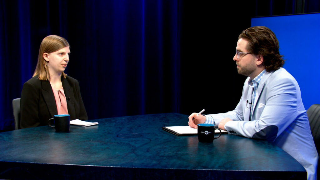 Two people sit across from each other at a table in a studio, both with notepads and coffee mugs, engaged in a serious conversation.