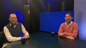 Two men sit at a table with black mugs in a studio setting, with a large blank blue screen and studio lights in the background.