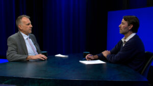 Two men sit across from each other at a round table in a studio, engaged in conversation. One has papers in front of him; both have coffee mugs. The background is dark blue.
