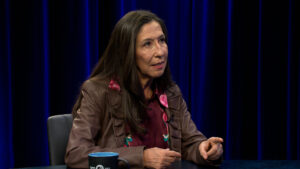 A woman with long brown hair, wearing a brown jacket and maroon blouse, sits at a table and gestures while speaking. Blue curtains hang in the background.