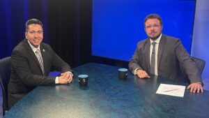 Two men in suits sit at a blue table with mugs and papers in a TV studio, facing the camera. A large blue screen is visible in the background.