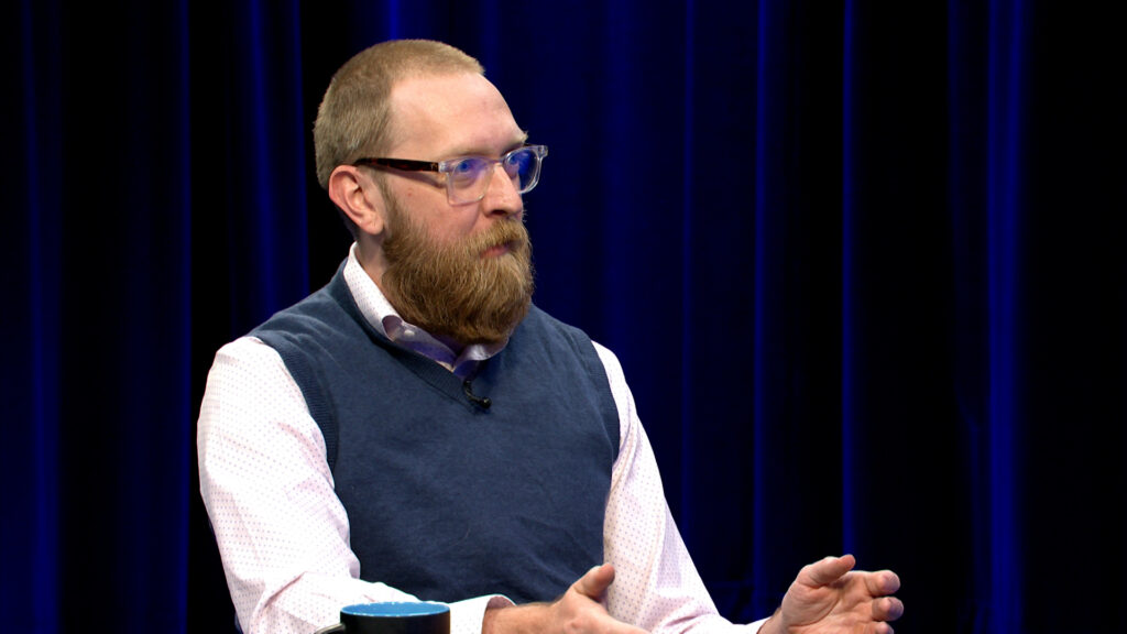 A man with a beard and glasses sits at a table, gesturing with his hands, against a dark blue curtain background.