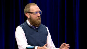 A man with a beard and glasses sits at a table, gesturing with his hands, against a dark blue curtain background.