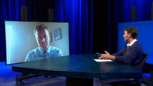 A man sits at a table in a studio, speaking to another man displayed on a large video screen during a remote interview.
