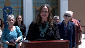 A woman speaks at a podium outdoors, surrounded by several people standing behind her.