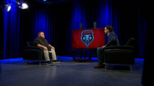 Two people sit in armchairs facing each other in a TV studio with blue lighting and a screen displaying a wolf logo and the word "LOBOS" between them.