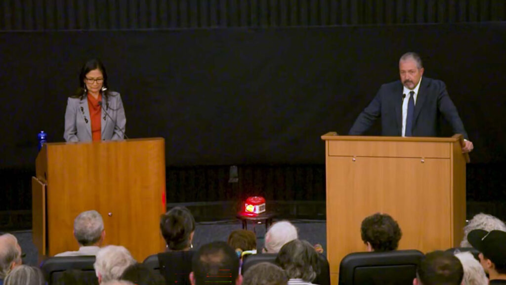 Two people stand at separate podiums facing an audience, appearing to participate in a debate or public discussion in an auditorium.