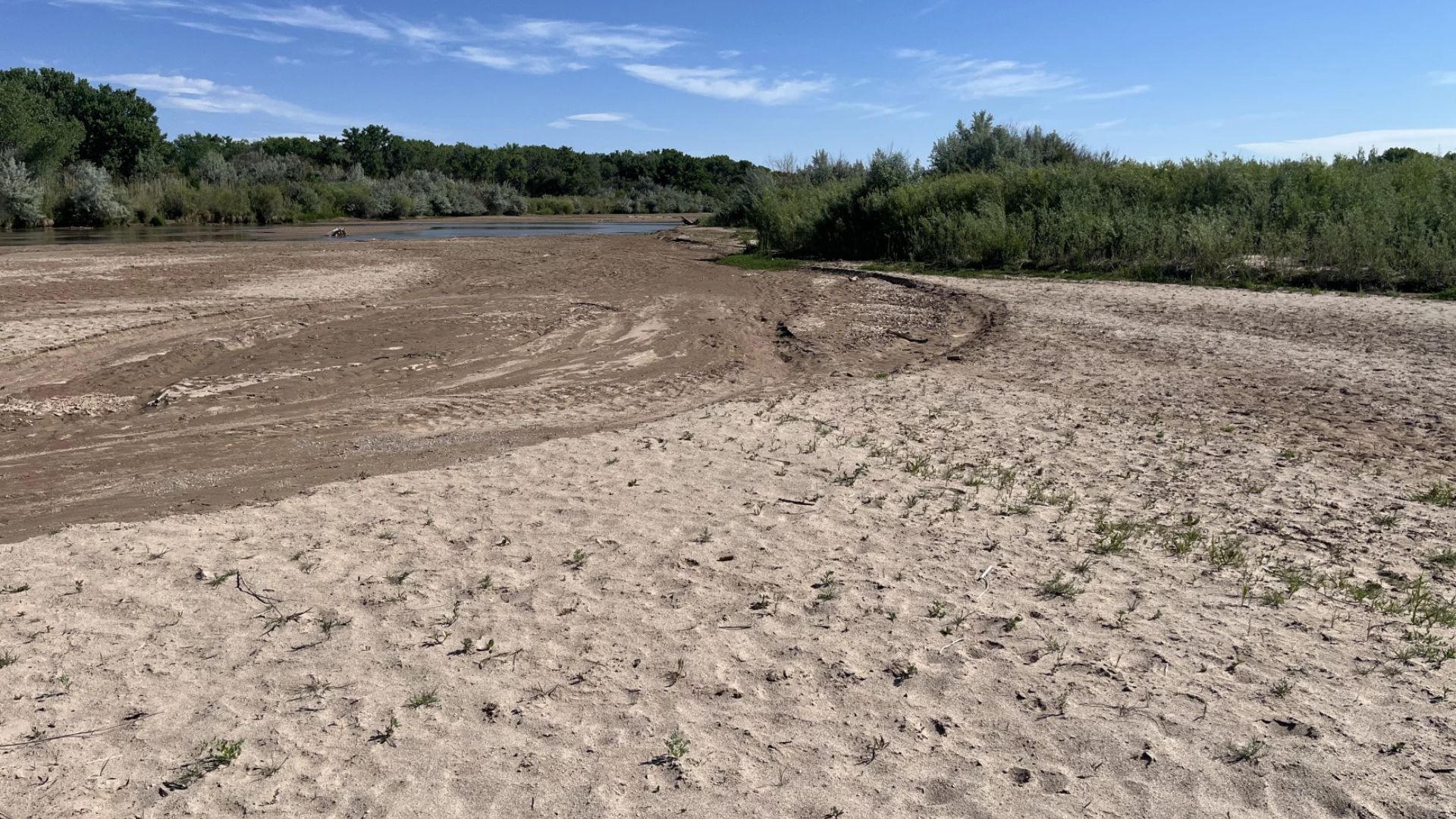 A sandy, dry riverbank with sparse vegetation leads to a small body of water under a clear blue sky with some clouds. Dense green trees line the background.