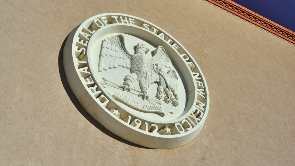 The Great Seal of the State of New Mexico, featuring an eagle and banner, is mounted on a tan wall with the year 1912 displayed at the bottom.