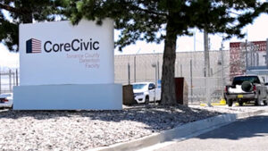 A sign for CoreCivic Torrance County Detention Facility stands by a road, with a fence, vehicles, and buildings visible in the background.