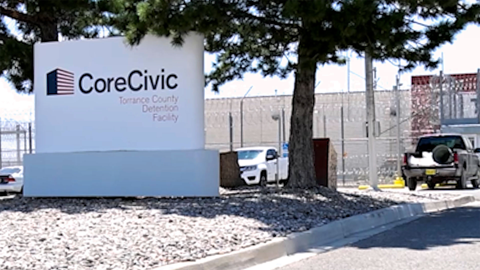 A sign for CoreCivic Torrance County Detention Facility stands by a road, with a fence, vehicles, and buildings visible in the background.