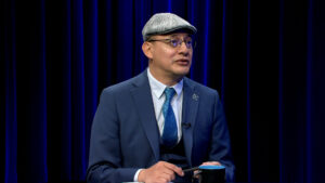 A man in a blue suit, tie, and flat cap sits in front of dark blue curtains, holding a pen and black mug while speaking.