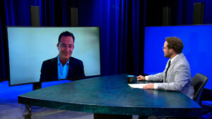 A man in a suit appears on a large monitor via video call, while another man in a suit sits at a desk facing the screen in a television studio.