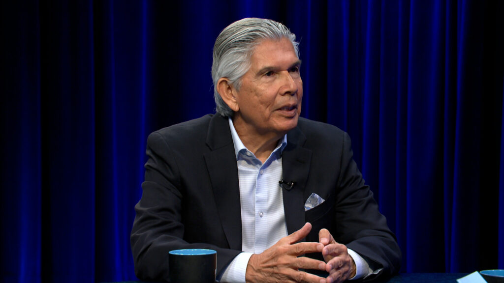 A man in a suit sits at a table, speaking and gesturing with his hands, with a blue curtain backdrop and a cup in front of him.