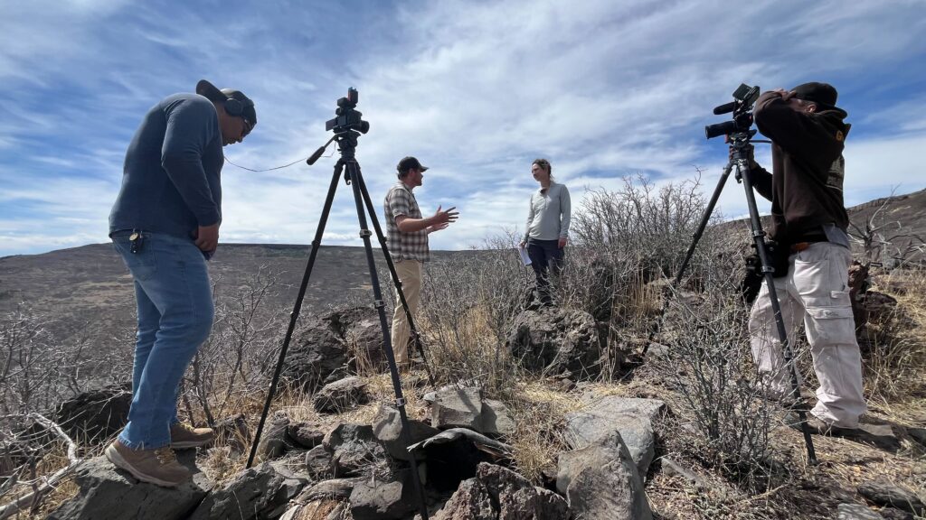 Two people talk on rocky terrain as two camera operators film them; all are outdoors under a partly cloudy sky.