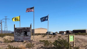 Three flags fly beside a sign reading “Forget Me Not Veteran’s Park, Glory Bound Baptist Church” in a desert area with buildings, cars, and power lines in the background.