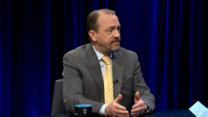 A man in a gray suit and yellow tie sits at a table, gesturing with his hands while speaking, with a blue curtain in the background.