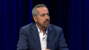 A man in a blue suit and white shirt sits at a table, speaking or listening, with a dark blue curtain background.