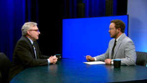 Two men sit across from each other at a table in a studio, engaged in conversation, with a blue screen in the background and coffee mugs on the table.