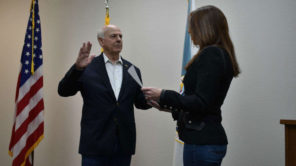 An older man raises his right hand while being sworn in by a woman holding papers, with American and state flags in the background.