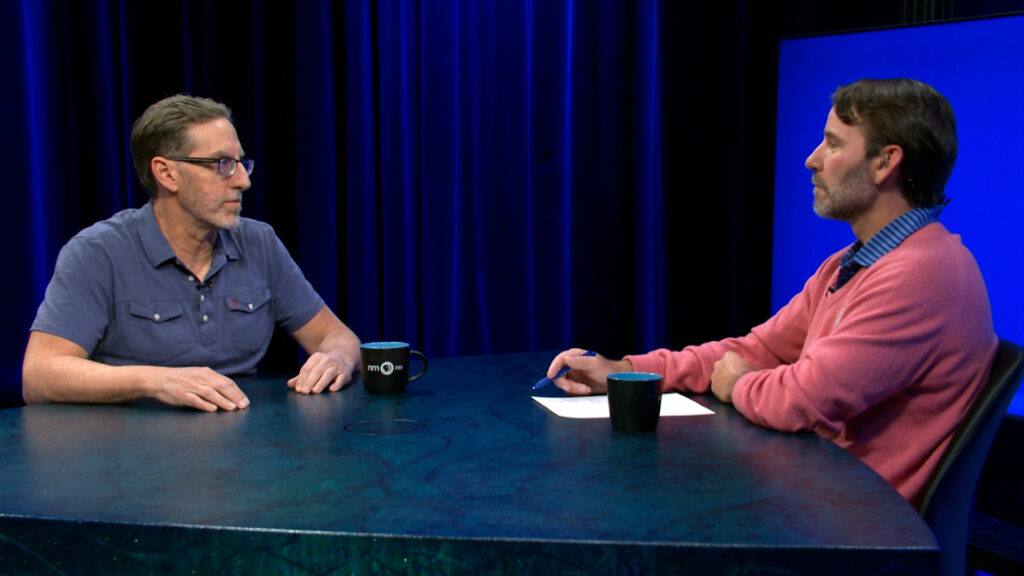 Two men sit across from each other at a round table with mugs and a notepad, talking in a studio with blue lighting and a dark background.