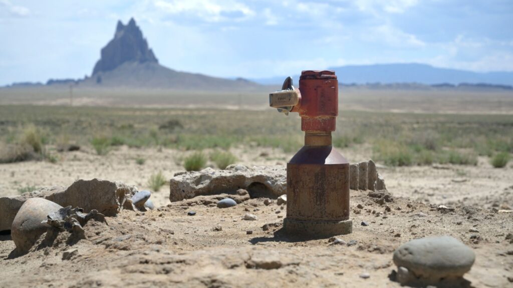 A rusty propane tank standing upright in a dry, rocky desert with a large rock formation in the background.