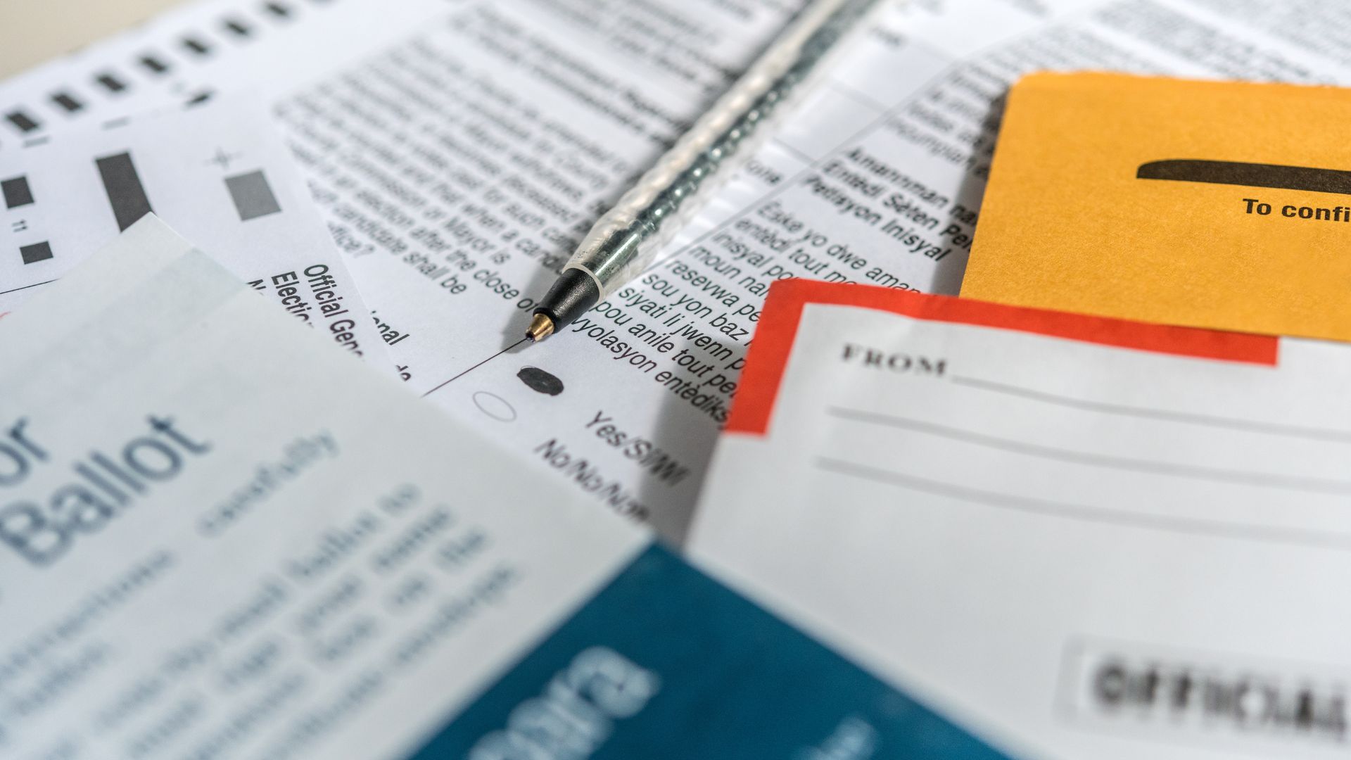 Close-up of a pen resting on a ballot paper, surrounded by election materials and envelopes, suggesting the process of voting by mail.