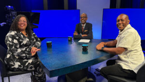 Three people sit around a blue table in a studio setting with blank blue screens in the background. Each has a cup in front of them and appears to be in conversation.