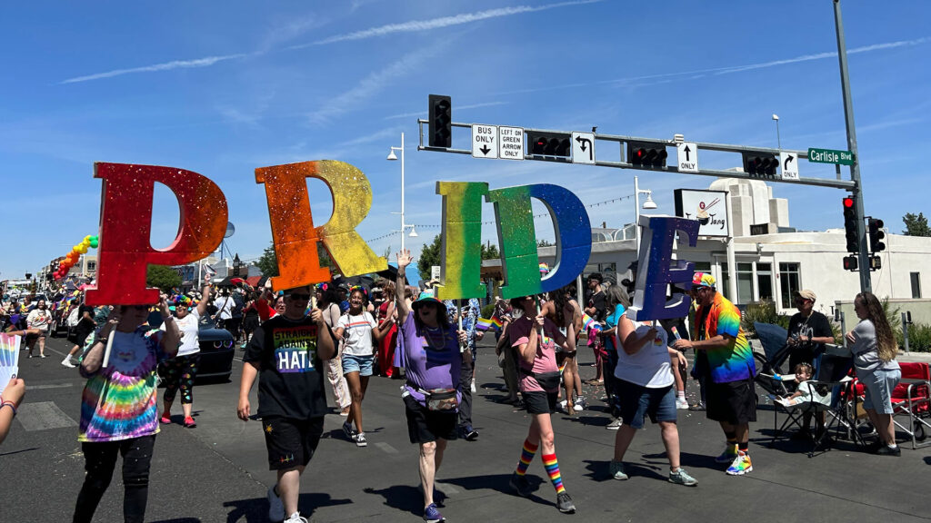 A group of people march in a parade holding large, colorful letters spelling "PRIDE" on a city street under a blue sky.