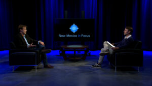 Two people sit in armchairs on a studio set with blue lighting, having a conversation for a TV program called "New Mexico in Focus," as displayed on the screen behind them.