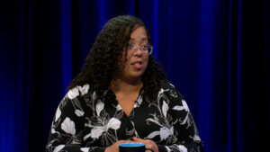 A person with curly hair and glasses, wearing a black and white floral shirt, sits at a table and speaks in front of a blue curtain backdrop.