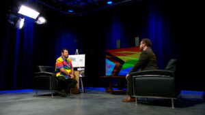Two people sit facing each other in a studio with stage lights, dark blue curtains, and a large LGBTQ+ pride flag displayed on an easel between them.