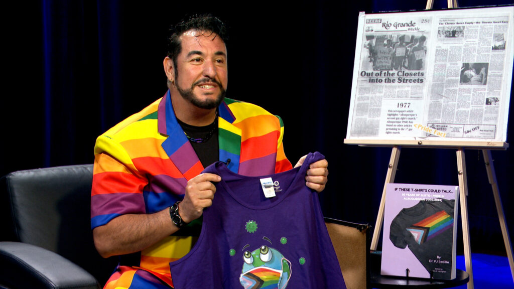 A man in a rainbow-striped jacket holds up a purple T-shirt with a rainbow graphic. Behind him are a newspaper article and a book displayed on easels.