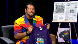 A man in a rainbow-striped jacket holds up a purple T-shirt with a rainbow graphic. Behind him are a newspaper article and a book displayed on easels.