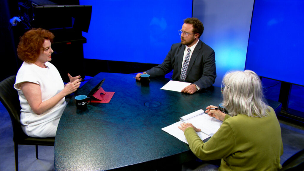 Three people sit around a table in a studio with blue screens, engaged in a discussion. Two have notepads and pens, while one gestures with hands. Cups and tablets are on the table.