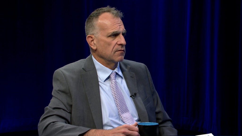A man in a gray suit and light purple tie sits holding a mug, with a serious expression, against a dark blue curtain backdrop.