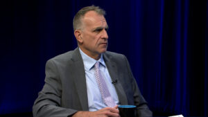 A man in a gray suit and light purple tie sits holding a mug, with a serious expression, against a dark blue curtain backdrop.