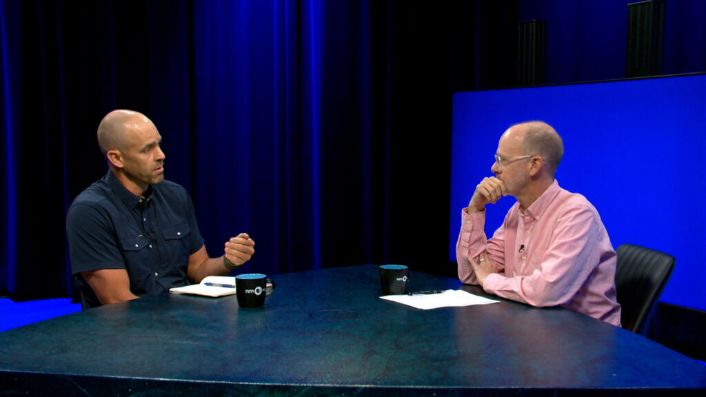 Two men sit across from each other at a table, engaged in conversation, with blue lighting and mugs on the table.