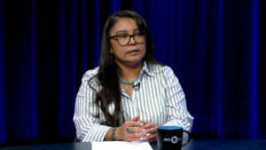 A woman with long dark hair and glasses, wearing a striped shirt, sits at a table with papers and a mug, speaking in front of a blue curtain.